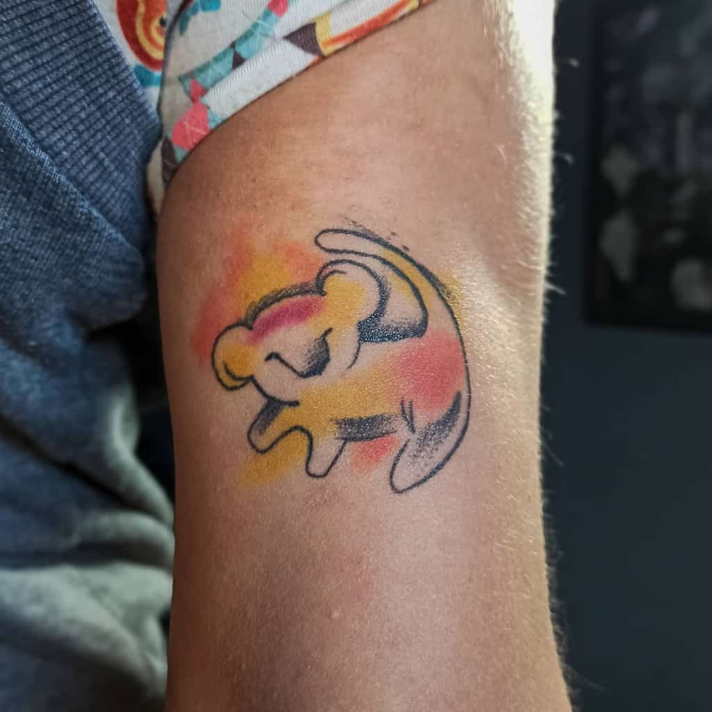 Minimalistic tattoo of Simba in an expressive pose, outlined with black ink and surrounded by soft watercolor splashes of yellow, red, and orange