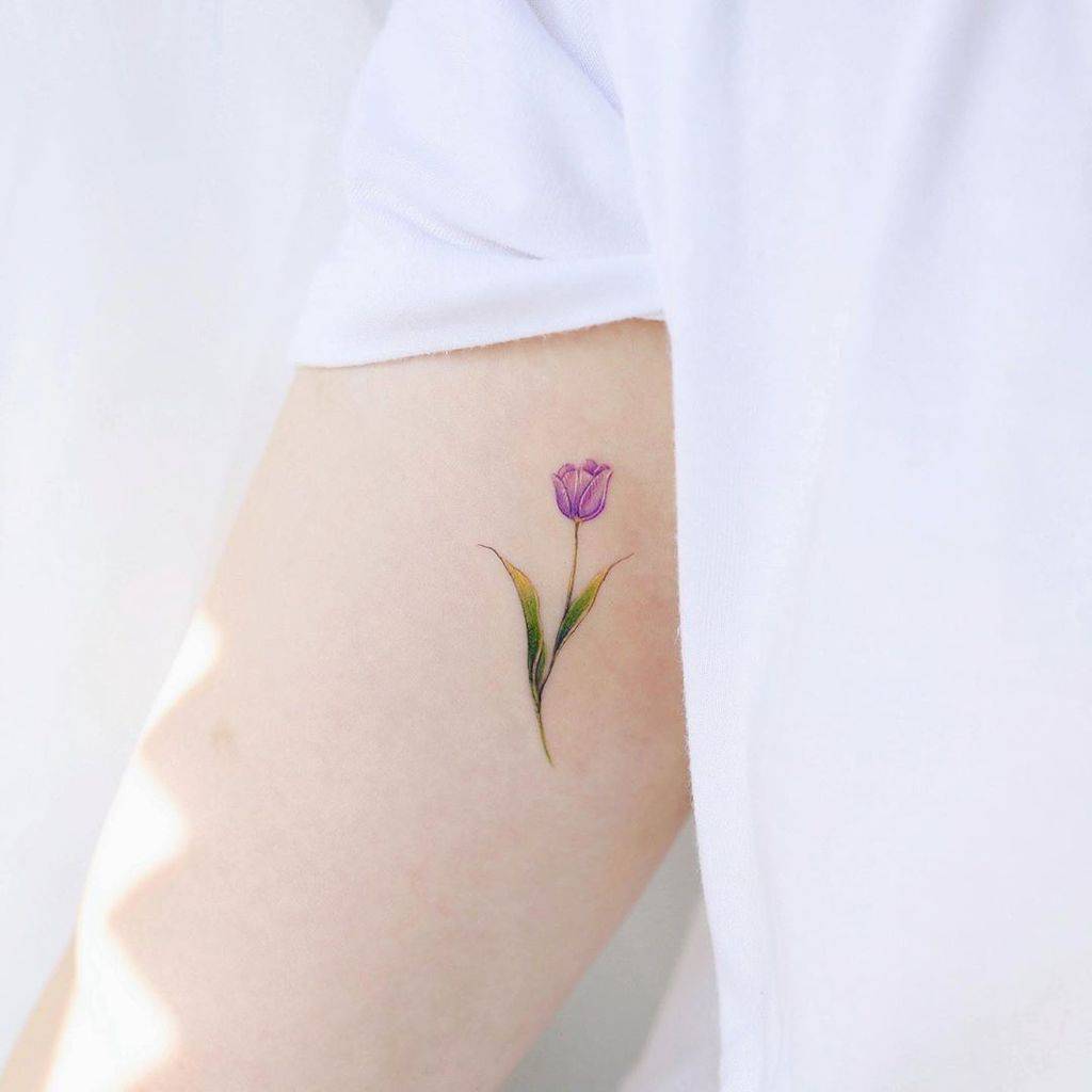 A small, delicate purple tulip tattoo on the upper arm, featuring fine details with soft green leaves in a realistic watercolor style.