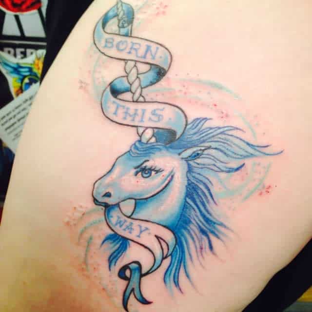 Tattoo of a blue unicorn with a banner that reads "Born This Way" wrapped around its horn