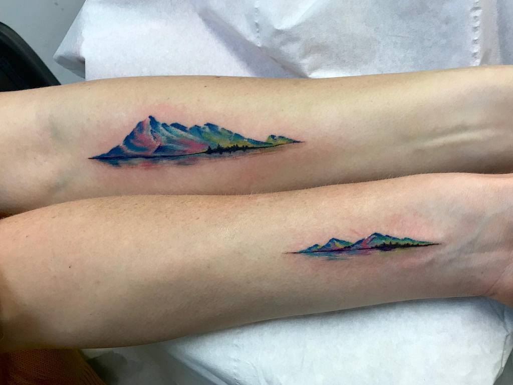 Matching colorful mountain landscape sister tattoos, featuring vibrant blues, greens, and purples