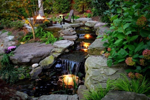 Backyard waterfall with illuminated stream, stone path, lush greenery, and cozy seating area.