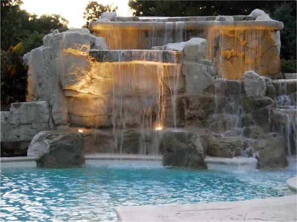 Backyard pool with illuminated cascading waterfalls over large rocks.
