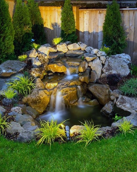 Small backyard waterfall with natural rocks, pond, and decorative lighting at night.