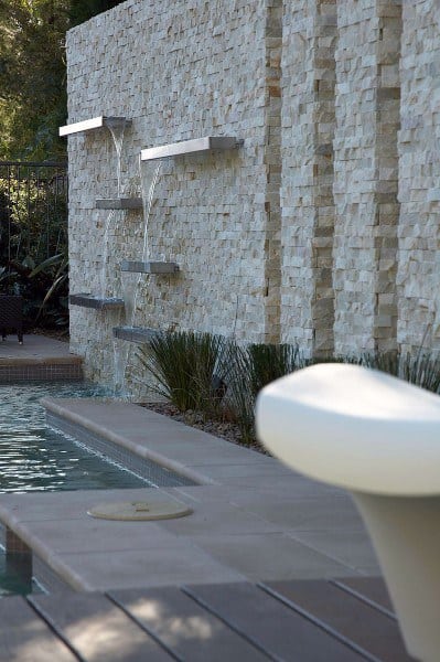 Layered wall-mounted steel waterfalls flowing into a modern pool with textured stone backdrop.