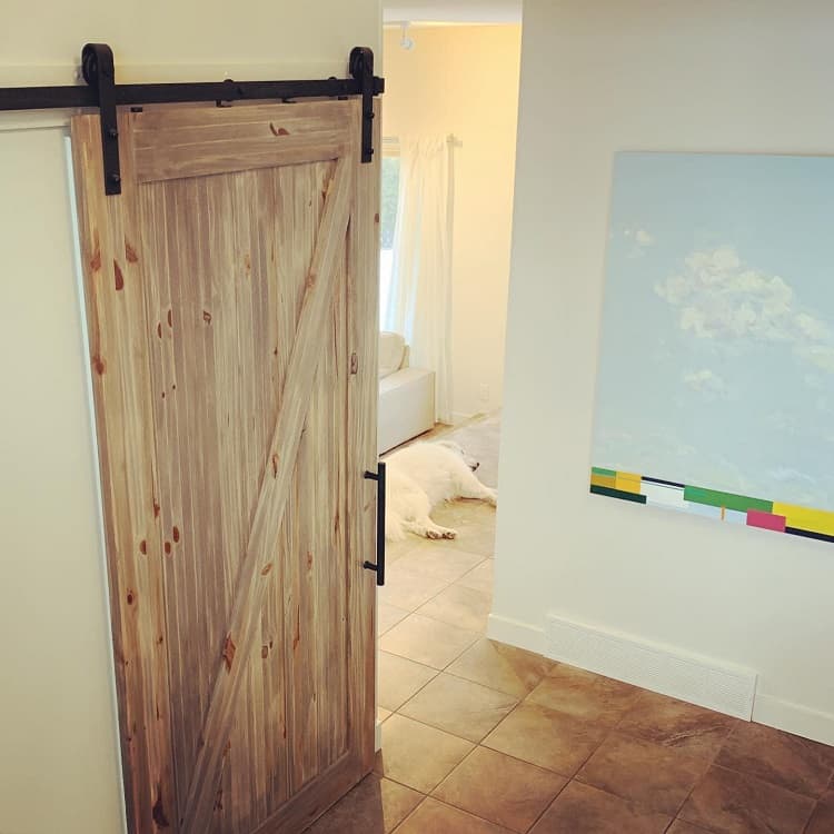 Weathered wood sliding barn door with black hardware leading to a cozy living space with tiled floors.