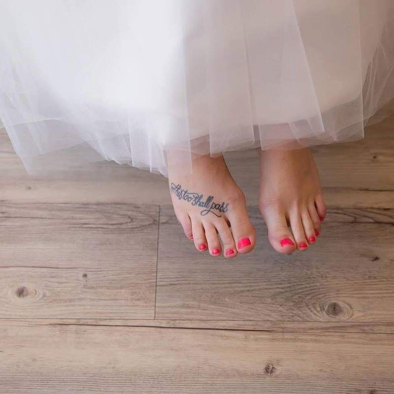 A "This Too Shall Pass" tattoo in cursive on the top of a person's foot, with pink toenail polish visible.