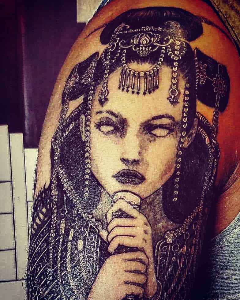 Dark-themed tattoo of a mysterious woman with white eyes, intricate headpiece, and ornate jewelry, blending gothic and fantasy elements.