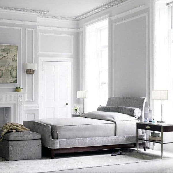 Elegant bedroom with a plush grey bed, white walls, large windows, nightstand, and a small ottoman