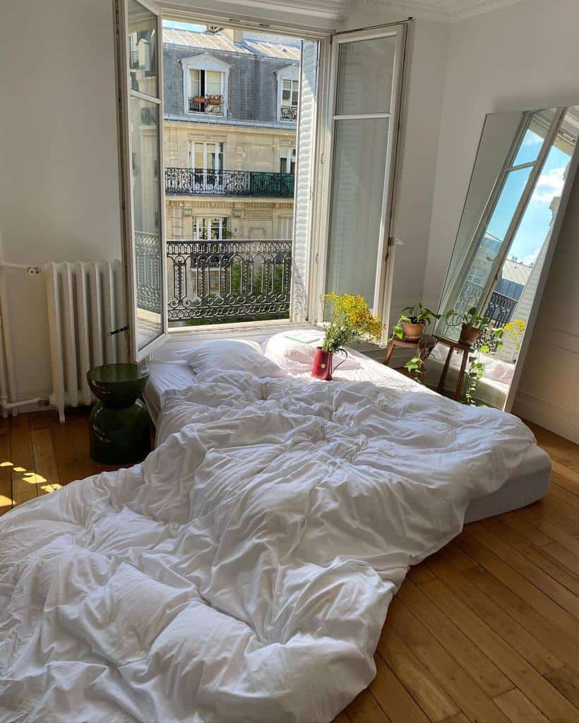 Cozy European-style bedroom with white bedding, open window, and wooden floors.