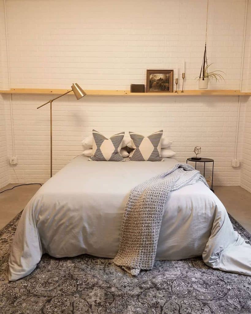 White basement bedroom with floating wooden shelf, geometric pillows, and cozy throw blanket.