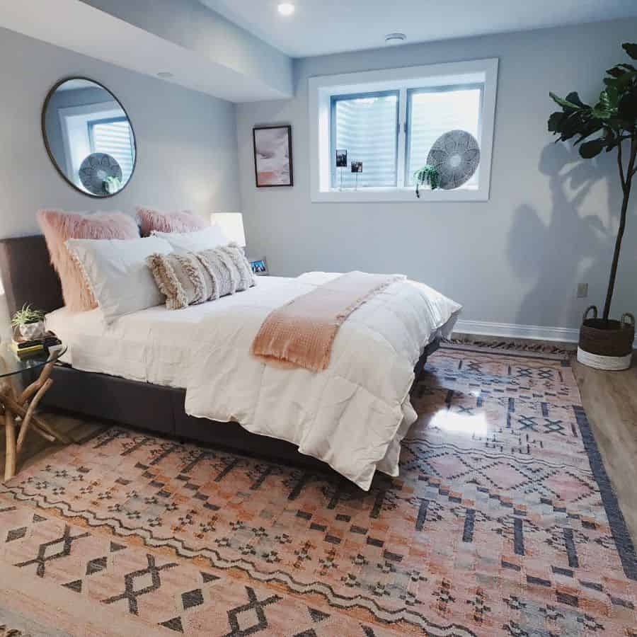 White basement bedroom with pink accents, patterned rug, and round mirror above the bed.