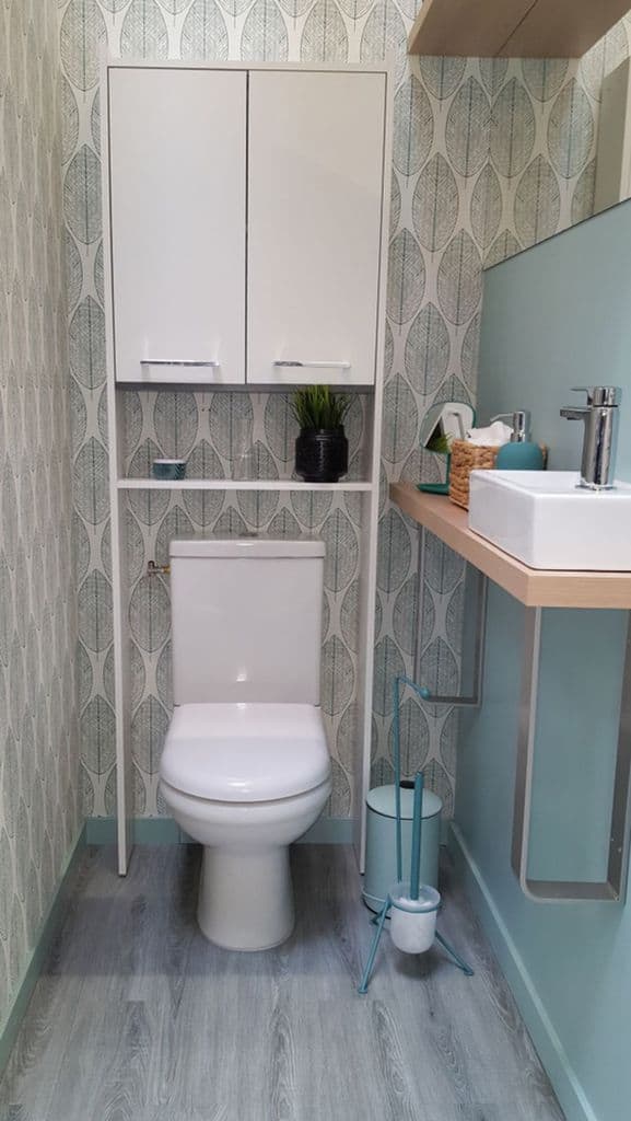 Small bathroom with leaf-patterned wallpaper, white fixtures, and light wood accents.