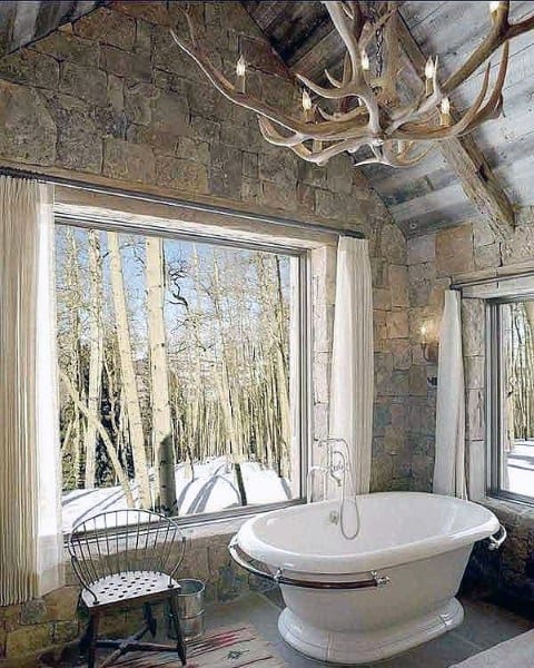Rustic bathroom featuring a freestanding tub, stone walls, antler chandelier, and snowy view