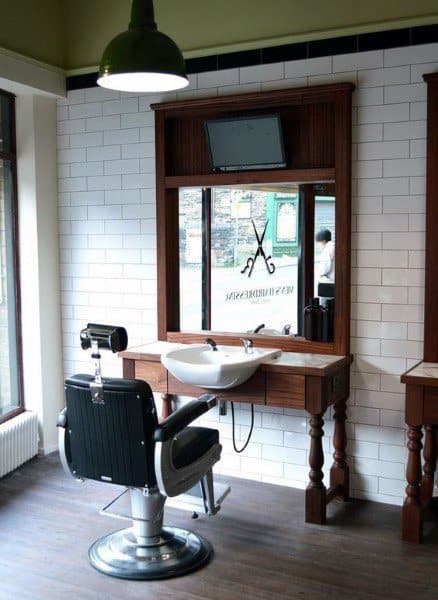 Barber station with white brick walls, wooden frame mirror, black chair, and washbasin.