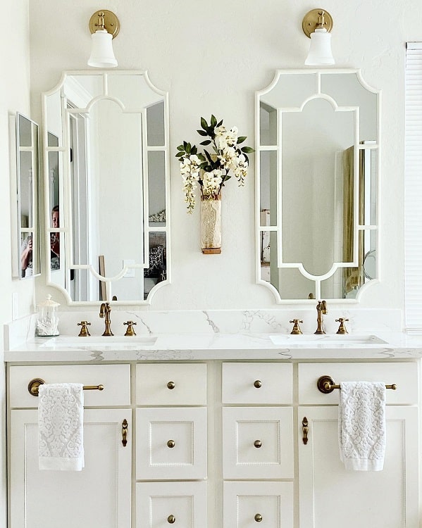 Farmhouse-style white bathroom with a double vanity, ornate mirrors, and gold fixtures featuring a flower arrangement on the wall