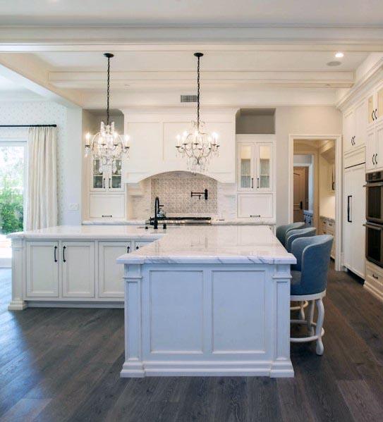 A sleek white kitchen with marble countertops, crystal chandeliers, and blue bar stools.