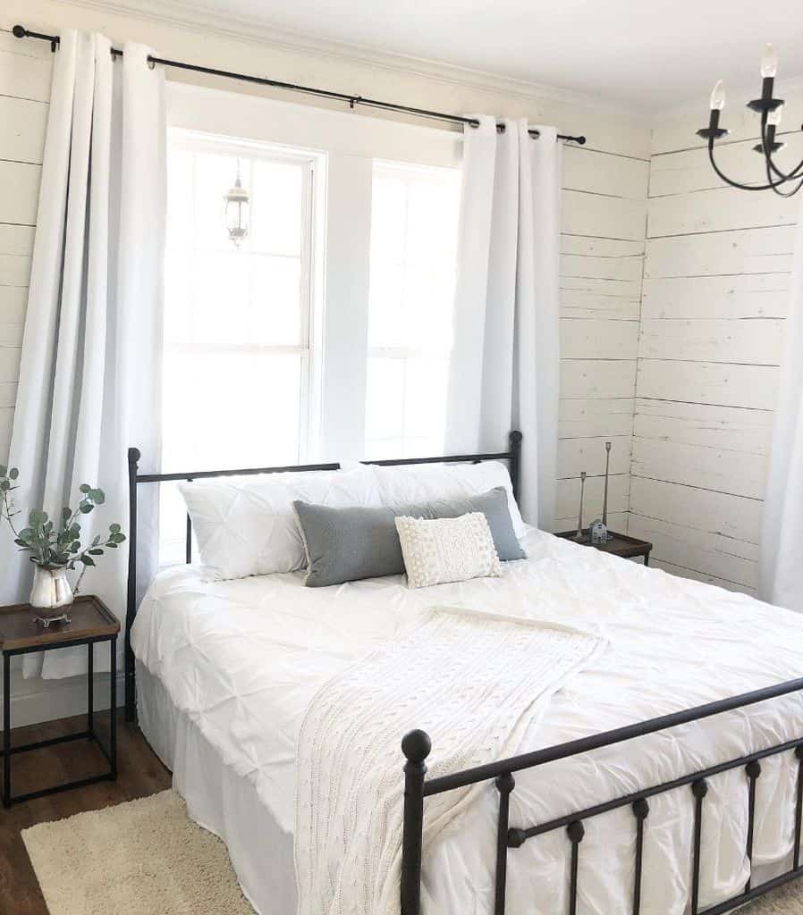 Farmhouse bedroom with white bedding, rustic shiplap walls, black metal bed frame, and a small plant on the bedside table