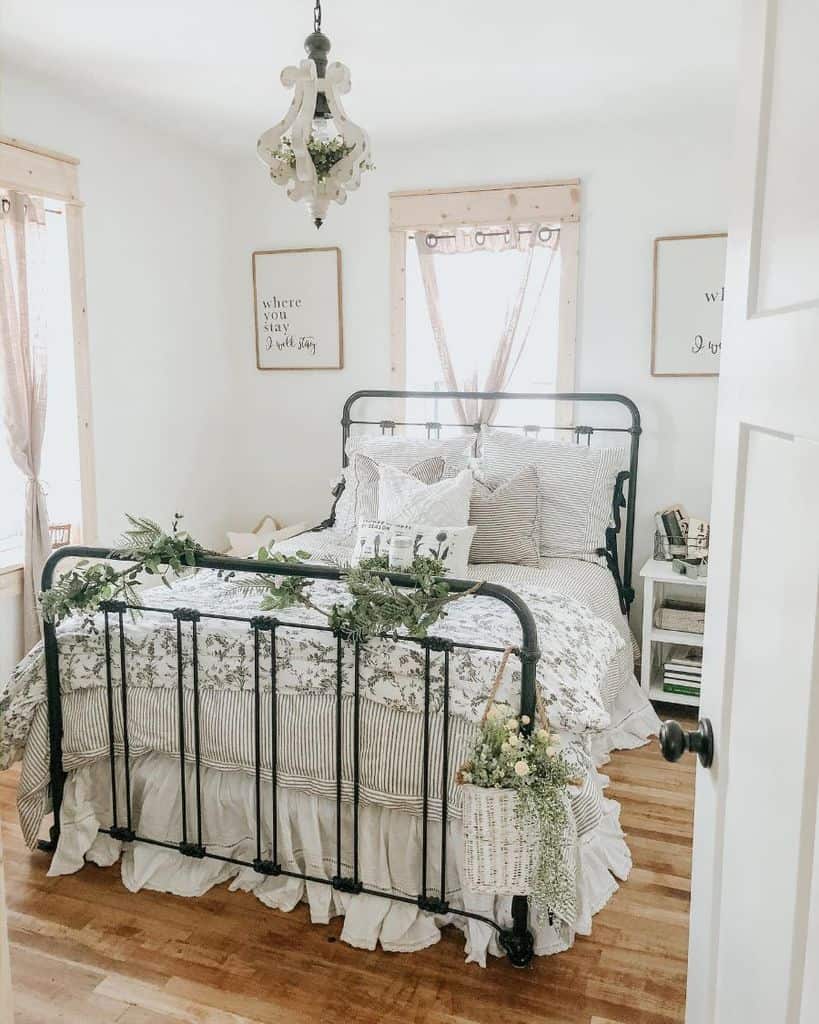 Cozy, rustic farmhouse bedroom with a metal bed, floral bedding, decorative greenery, and light-filled windows