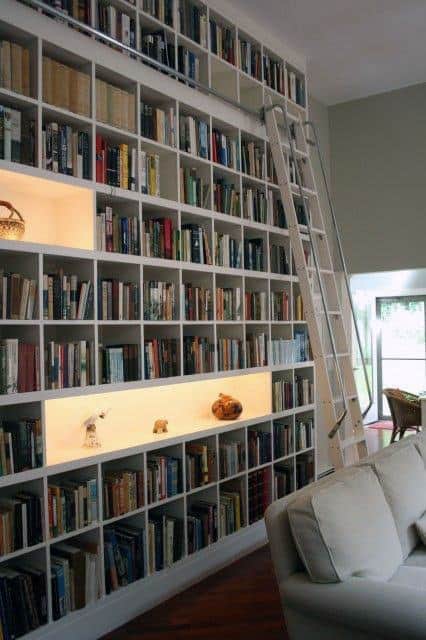 A tall bookshelf filled with books and decorative items, with a ladder and a white couch in the foreground
