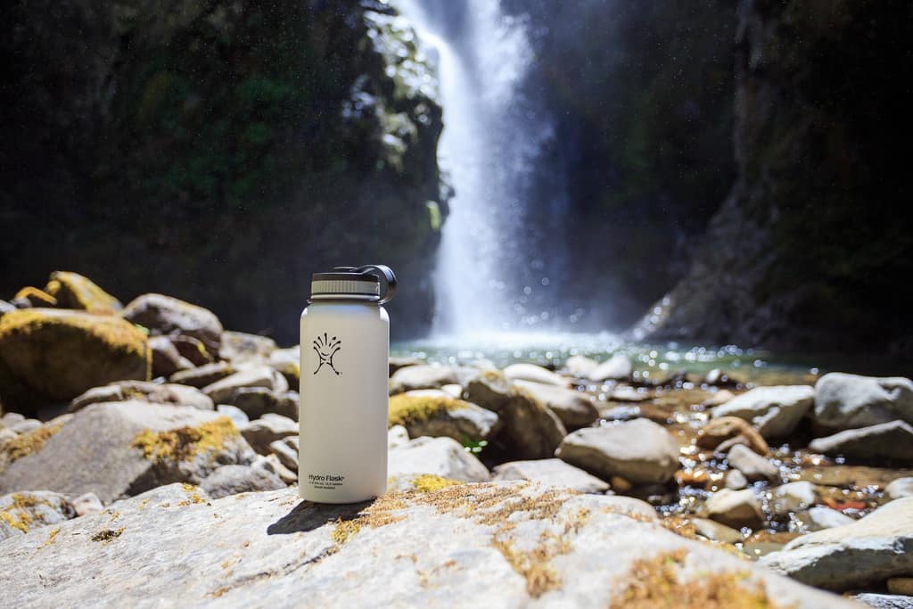 white hydroflask with company logo sitting on rock by a waterfall surrounded by tall cliffs