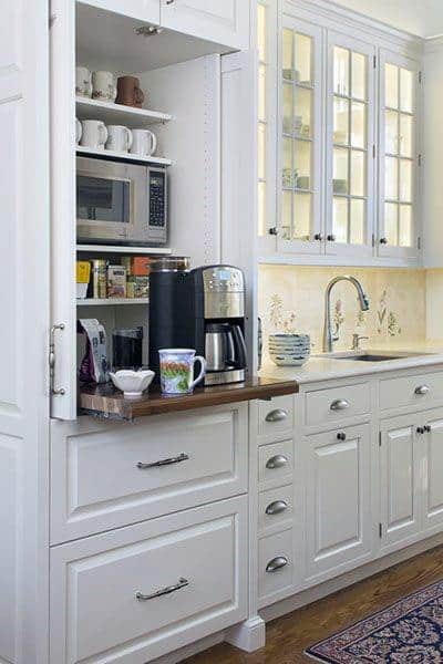 White kitchen coffee station with pull-out shelf, microwave, mugs, and built-in storage.