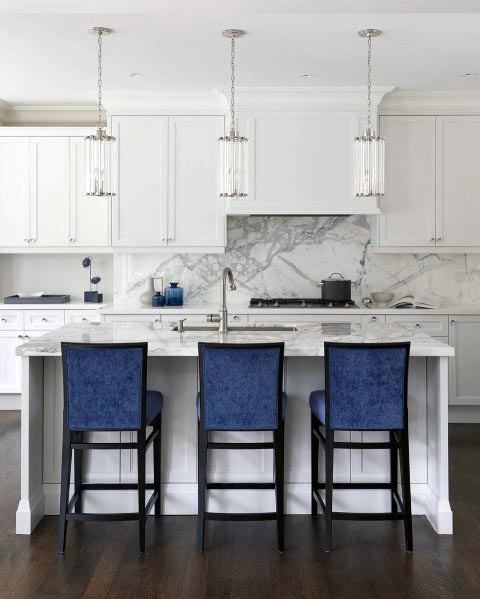 White kitchen with marble backsplash, navy blue bar stools, and pendant lighting.