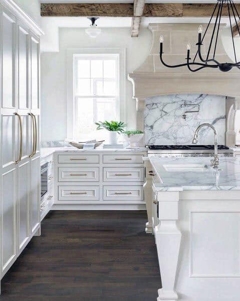 White kitchen with marble backsplash, exposed beams, gold hardware, and black chandelier.