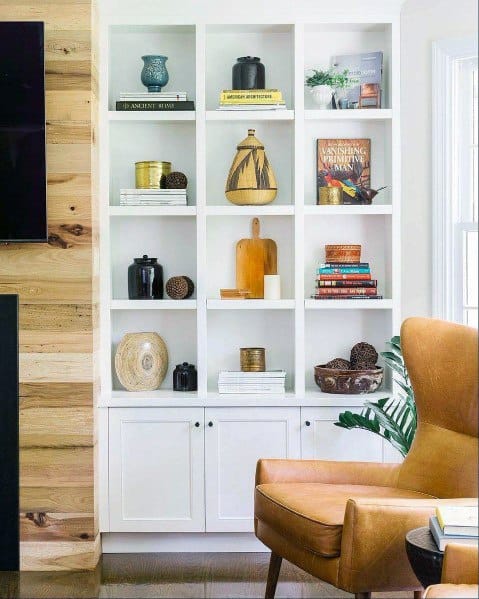 A bright and stylish built-in bookcase with white painted shelves and cabinets, featuring a curated mix of books, decorative pottery, and modern accents. Perfectly paired with a warm leather chair for a cozy reading corner.
