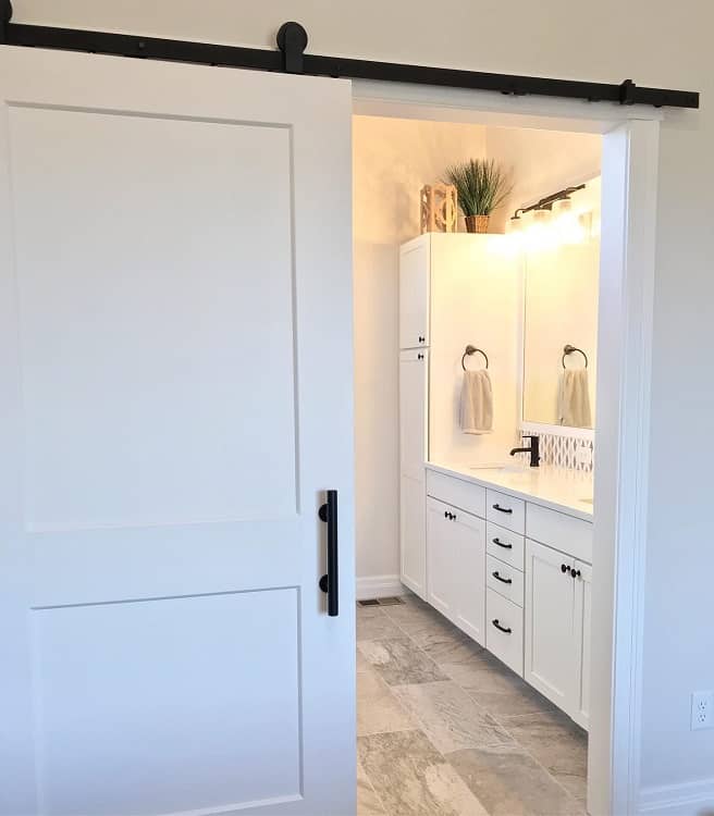Simple white sliding barn door leading to a bright bathroom with white cabinetry and tiled flooring.