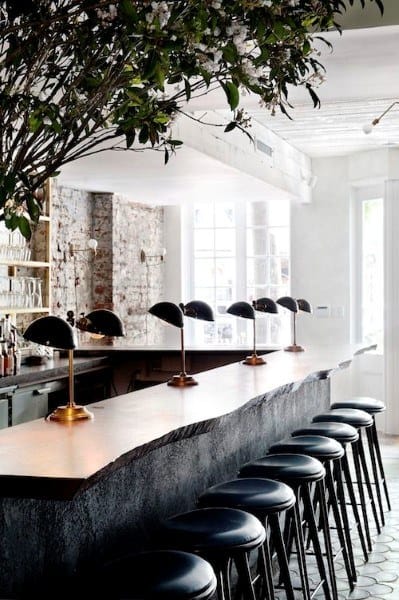 White stone bar top with modern black bar stools, gold lamps, and exposed brick wall backdrop.