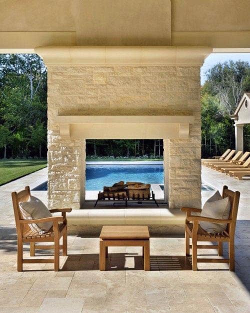Outdoor patio with wooden chairs and table, stone fireplace, pool in background, surrounded by greenery