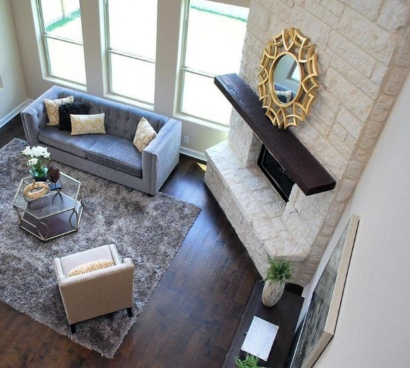Bright living room with white stone corner fireplace, dark wood mantel, gray sofa, and gold mirror.