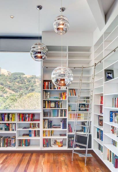 Modern library with tall white bookshelves, a ladder, and three pendant lights