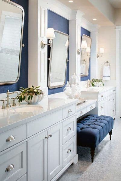 Bathroom with white double vanity, navy wall accents, large mirrors, and tufted blue bench.