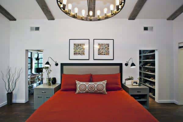 Bedroom with a vibrant red bedspread, two framed artworks, and a large chandelier; open closets flank each side