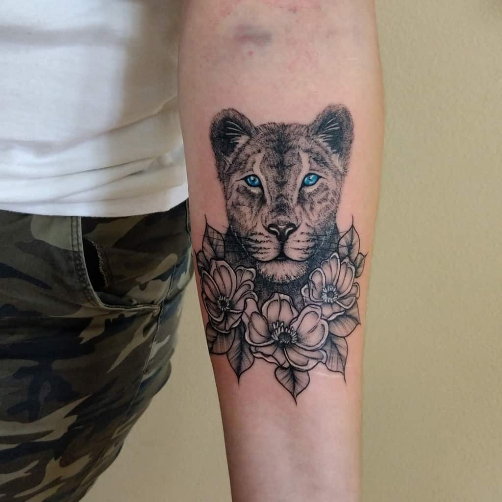Black and grey lioness forearm tattoo with vibrant blue eyes, surrounded by detailed flowers for a bold yet elegant design.