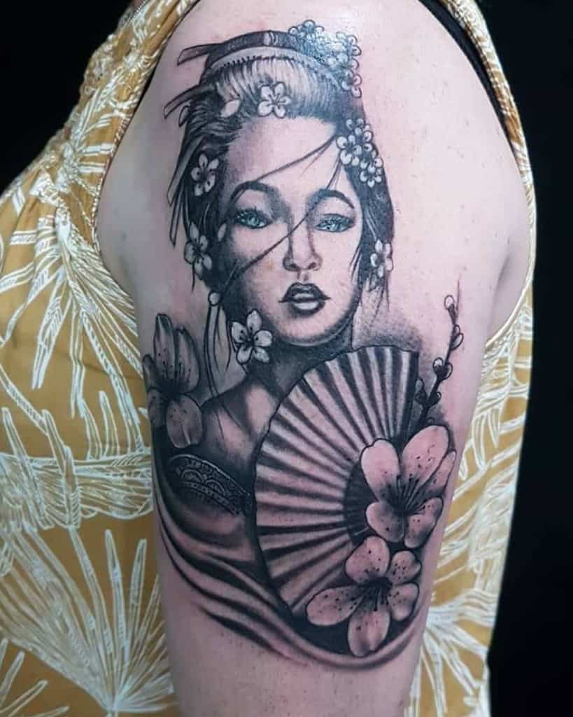 Black and gray upper arm tattoo of a Geisha with flowing hair, floral ornaments, a delicate fan, and cherry blossoms surrounding the design.