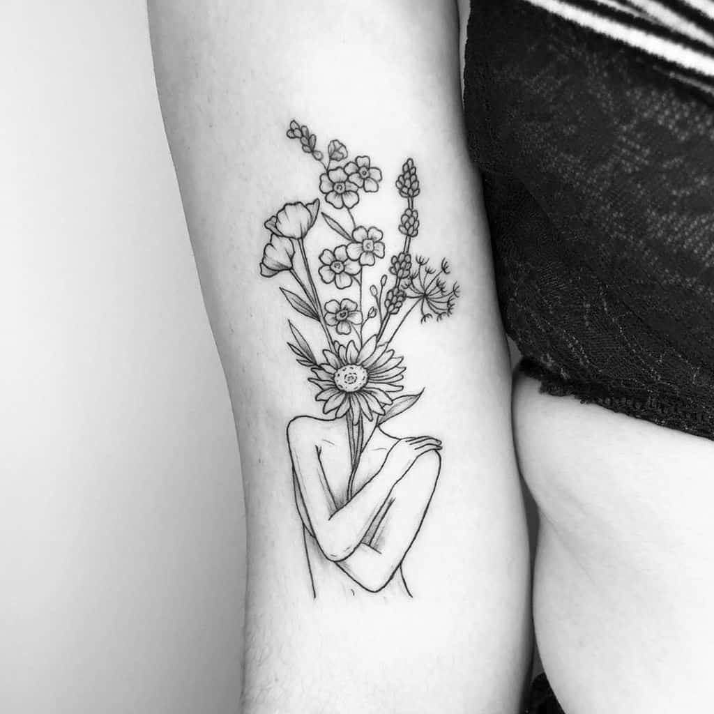 A tattoo on the upper arm featuring a human figure with its head replaced by a bouquet of various flowers, including daisies and poppies. The figure is crossing its arms over its body, creating a contrast between the delicate flowers and the abstract human form. The design is in black ink with fine detailing.