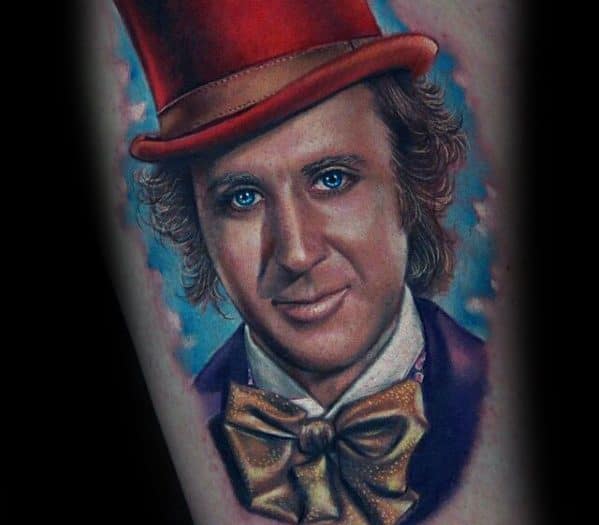 Realistic Willy Wonka tattoo with striking blue eyes, a red top hat, gold bow tie, and a dreamy blue cloud-like background.