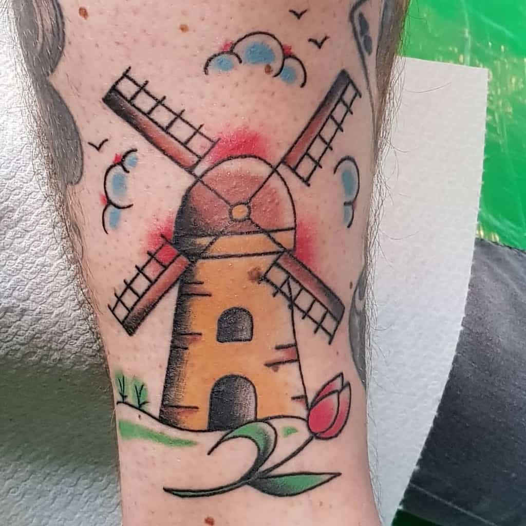 A colorful windmill tattoo on the leg, featuring a traditional Dutch windmill with a red tulip, green grass, and blue clouds in a bold style.
