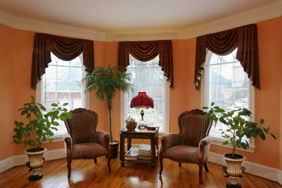 Traditional sitting area with patterned drapes, vintage chairs, red lamp, and indoor plants.