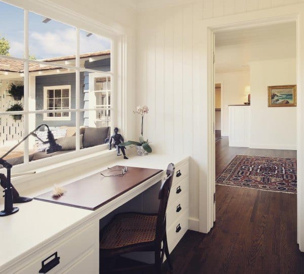 Bright home office with a built-in wooden desk, chair, and large window overlooking a patio; rug visible in the hallway