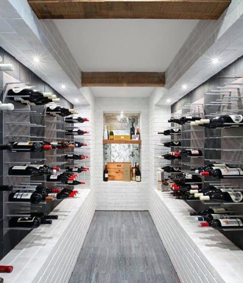 A narrow wine cellar with bottles on wall racks, wooden ceiling beams, and wine shelves at the end