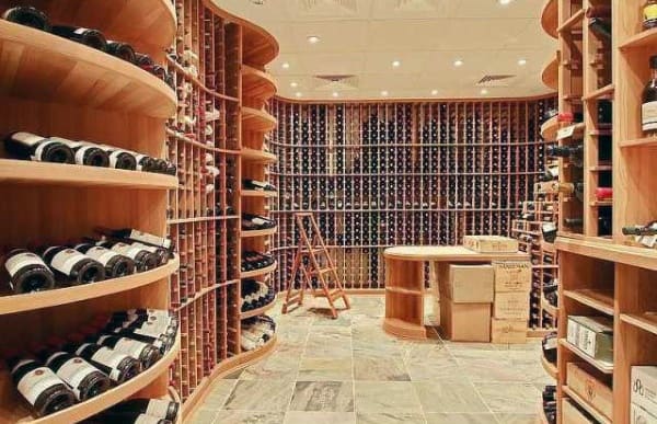 A wine cellar with wooden shelves lined with wine bottles and a wooden ladder in the center beckons wine enthusiasts