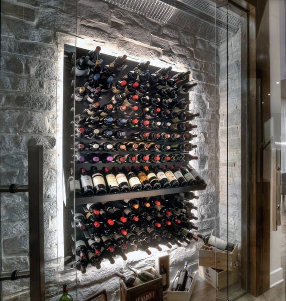 A modern wine rack with rows of fine wine and bottles against a stone wall, illuminated from behind