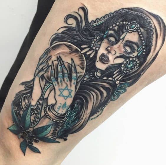 Intricate tattoo of a mystical gypsy woman with glowing eyes, ornate jewelry, and a crystal ball, featuring dark and teal shading on the arm.