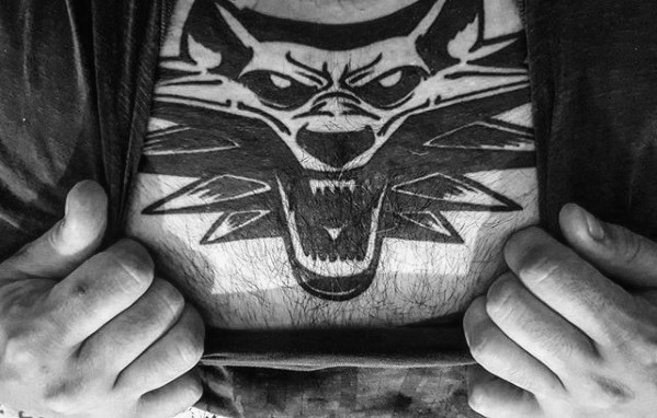 Bold black chest tattoo of the Witcher wolf medallion with sharp angular lines and a fierce expression, covering the upper torso.