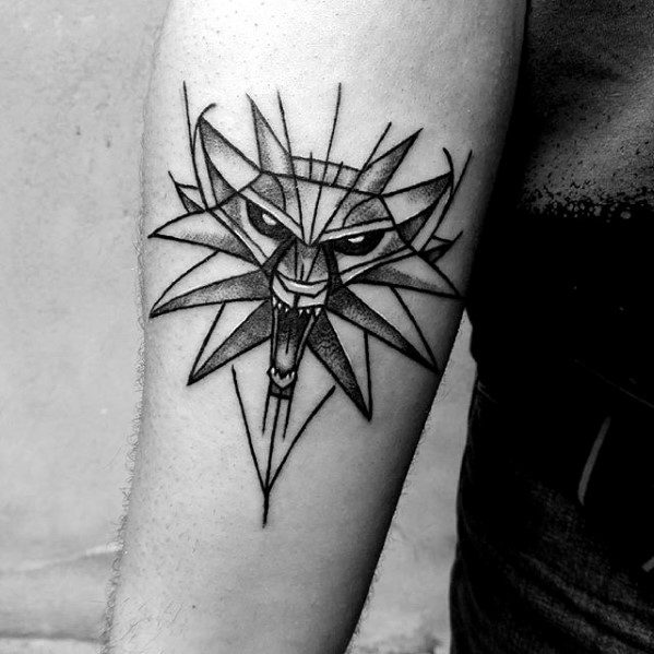 Minimalist Witcher tattoo on the upper arm featuring the wolf medallion in a geometric style with sharp lines and bold black ink.