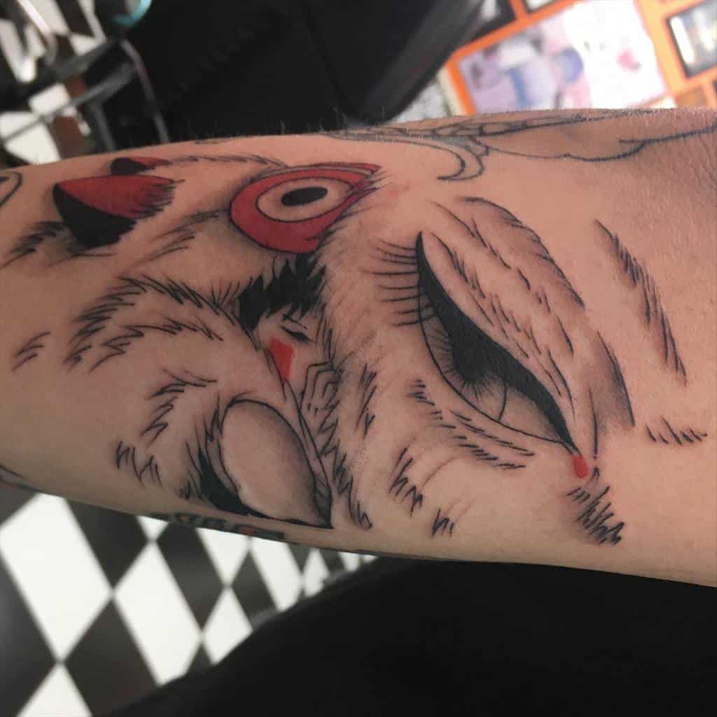 A tattoo on the forearm featuring a detailed wolf's face with a red-eyed headdress. The design incorporates delicate lines and shading, showing elements of the wolf's fur and the character's face.