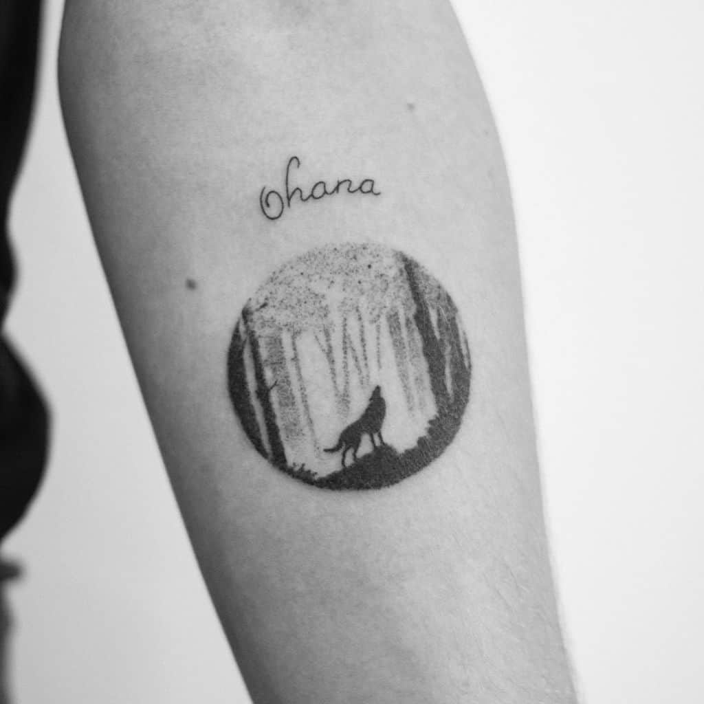 Minimalist tattoo of a wolf howling in a forest with 'Ohana' written above, symbolizing family, strength, and connection to nature.